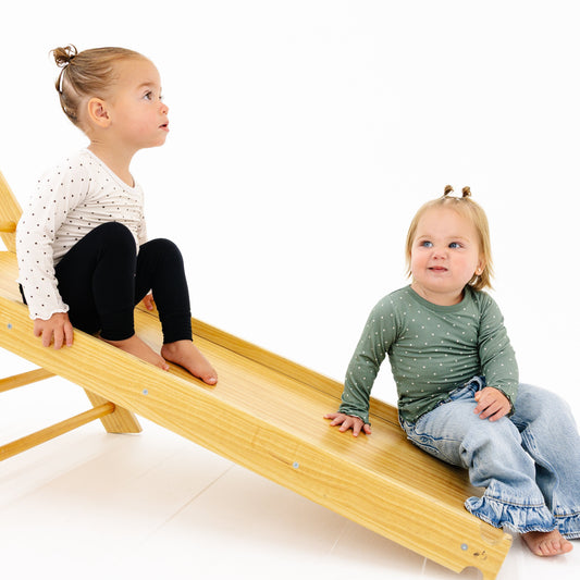 Two toddlers play on a light natural wood climbing slide against a seamless white background. The child on the left sits at the top wearing a white long-sleeve top with black polka dots and black pants, while the child on the right slides down wearing a green long-sleeve top and blue jeans.