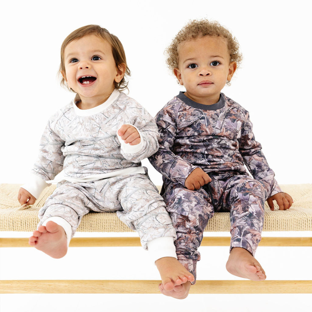 Two toddlers sitting on a wooden bench wearing matching patterned long-sleeve pajamas, photographed against a clean white studio background.