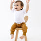 Child sitting on a small wooden stool in brown baby joggers with arms raised in a white pocket tshirt against a white background