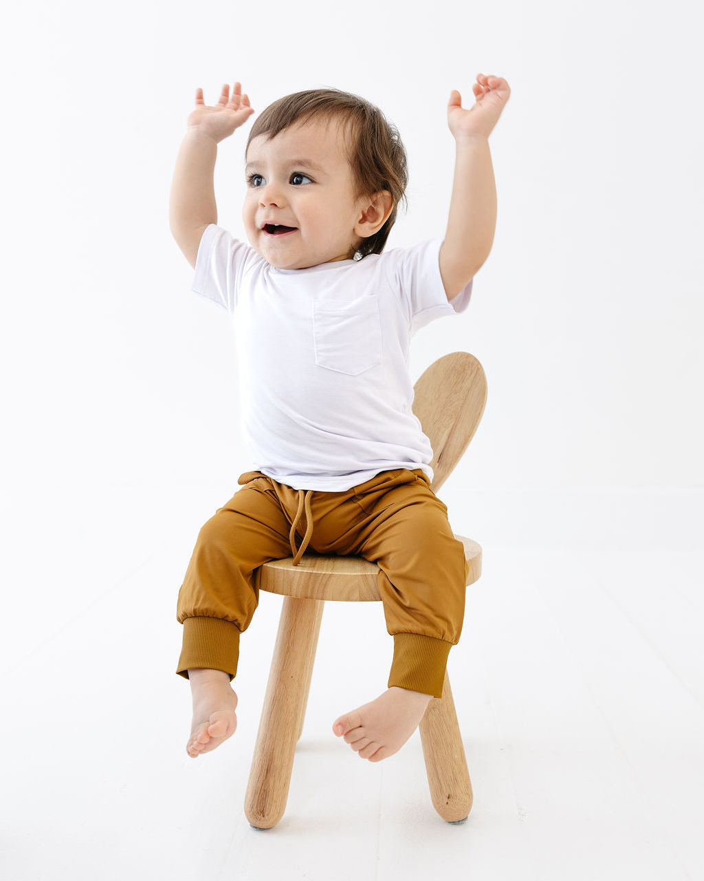 Child sitting on a small wooden stool in brown baby joggers with arms raised in a white pocket tshirt against a white background
